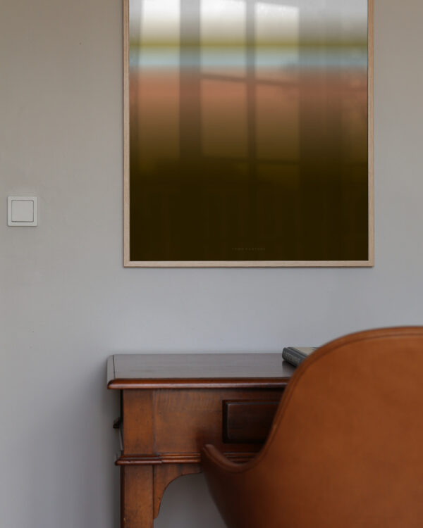 An oak frame with the art print inside called; Dusk 01, hanging on a white wall with a work table and chair below.