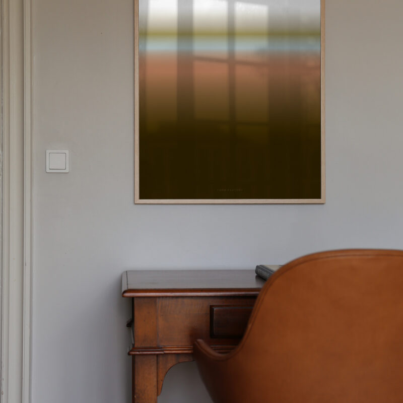 An oak frame with the art print inside called; Dusk 01, hanging on a white wall with a work table and chair below.